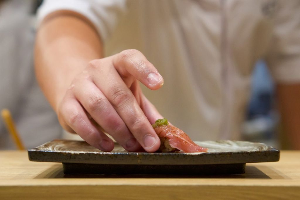 A skilled chef at Sushi Sora elegantly presenting a freshly made sushi dish, delicately placing it on a sleek plate against the backdrop of the restaurant's inviting and professional kitchen.